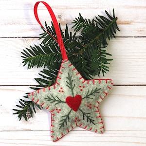 May include: A gray felt star ornament with a red felt heart and green felt pine branches embroidered on it. The ornament has a red ribbon for hanging.