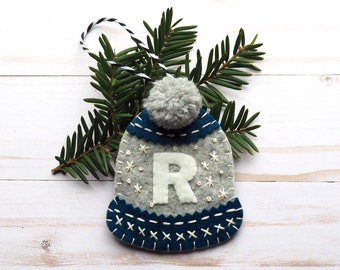 Pode incluir: Um enfeite de Natal de feltro em forma de chapéu de inverno azul e cinza com um pompom branco no topo. O chapéu tem uma letra "R" de feltro branco bordada na frente.