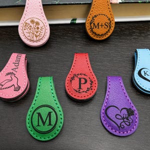 May include: A collection of seven teardrop-shaped magnetic bookmarks in various colors including pink, orange, red, green, purple, and light blue. Each bookmark features a unique black laser-engraved design, such as floral patterns, initials, names, and hearts.