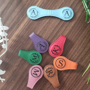 May include: A collection of seven leather magnetic clips in various colors including light blue, purple, red, green, orange, pink, and brown. Each clip features a black printed letter inside a circular floral wreath design for personalization.