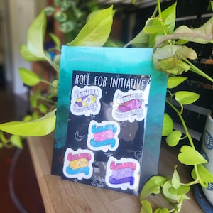 May include: A black sticker sheet with five colorful stickers that read "Roll for Initiative" and "Neurodivergency". The stickers feature a D20 die and are set against a teal and blue background. The image is framed by green foliage.