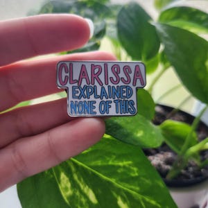 May include: A white enamel pin with a blue and pink border. The pin says "Clarissa Explained None of This".