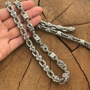 May include: A silver chain necklace with intricate designs and cross accents. The chain is made of linked oval shapes with detailed engravings.