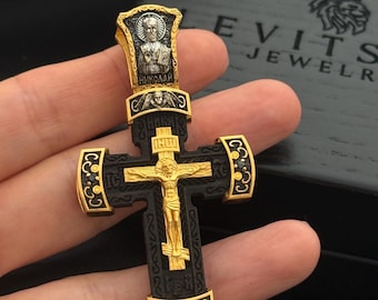 Pode incluir: Um pingente de cruz preto e dourado com uma pequena imagem de um santo no topo. A cruz é decorada com pedras pretas e as palavras "НИКОЛАЙ" estão inscritas no topo.