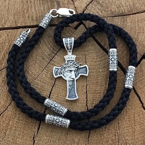 May include: A silver Jesus Christ pendant with a crown of thorns and the inscription "INRI" on a black braided cord necklace. The necklace has silver beads with a cross design.