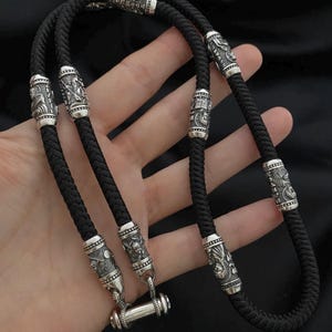 Op de afbeelding: Zwarte gevlochten ketting met sierlijke zilverkleurige cilindrische kralen. De kralen hebben gedetailleerde reliëfs van figuren en patronen. De ketting wordt getoond tegen een donkere achtergrond.
