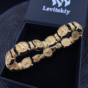Op de afbeelding: Een gouden armband met een zwart leren bandje en gouden medaillons met religieuze afbeeldingen. De armband zit in een zwarte doos met het woord "Levitskiy" op de deksel.
