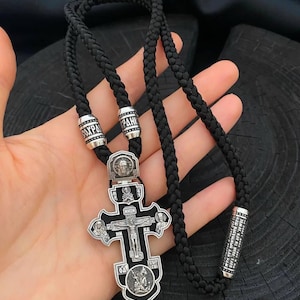 May include: A silver Orthodox cross pendant with black enamel and multiple religious figures. The cross is attached to a black braided cord with silver beads.