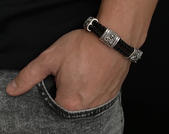 May include: A black leather bracelet with silver-toned metal accents. The bracelet features a braided leather strap and multiple decorative metal beads.