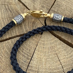 Puede incluir: Un collar de cordón trenzado negro con tapas de extremo doradas y plateadas. Las tapas de extremo tienen un diseño intrincado y el texto "ELIH" es visible en una de las tapas.
