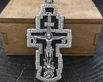 May include: A silver Orthodox cross pendant with black enamel and intricate details. The cross features a depiction of Jesus Christ on the front and Cyrillic text on the back.