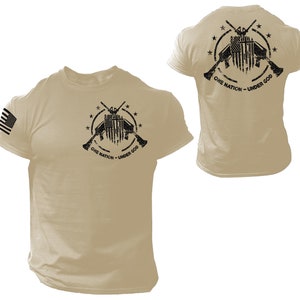 May include: Tan short-sleeved t-shirt with a black and white graphic of crossed rifles, an American flag, and the text "One Nation - Under God". The shirt has a small American flag on the left sleeve.