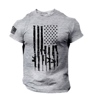 May include: A grey t-shirt with a distressed American flag design. The flag is made up of black guns instead of red and white stripes.