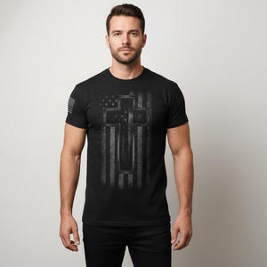May include: Black crew neck t-shirt with a distressed American flag and cross design in grey tones. A small American flag is on the left sleeve. The shirt is a classic fit.