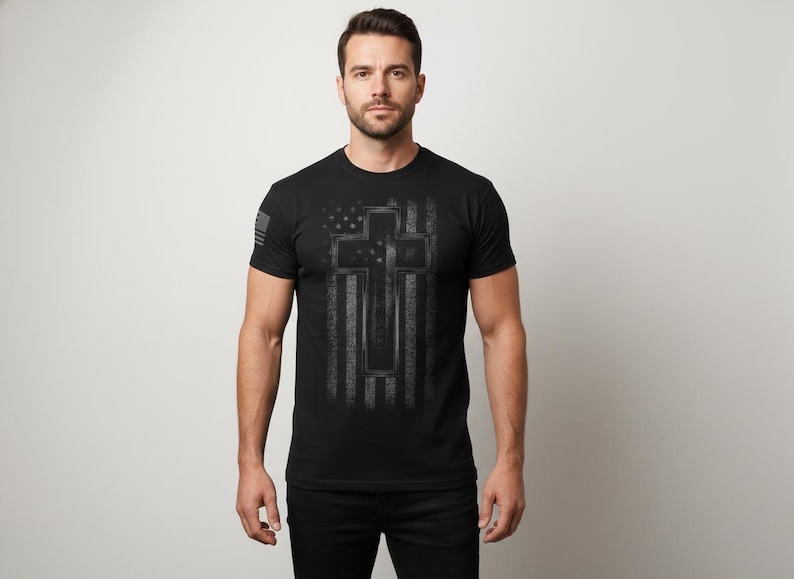 May include: Black crew neck t-shirt with a distressed American flag and cross design in grey tones. A small American flag is on the left sleeve. The shirt is a classic fit.
