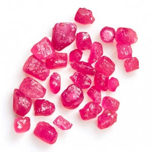 Puede incluir: Una colección de piedras preciosas de rubí rosa, algunas con bordes ásperos, esparcidas sobre un fondo blanco.