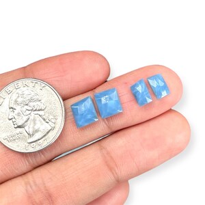 Può includere: Quattro cabochon di gemme sfaccettate blu chiaro di varie dimensioni sono esposti accanto a una moneta. Le gemme hanno una forma quadrata o rettangolare con un aspetto leggermente traslucido. Le gemme sono tenute nella mano di una persona.