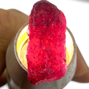 Puede incluir: Una piedra preciosa roja grande, áspera, con un color profundo y rico. La piedra preciosa tiene una forma irregular y una apariencia ligeramente translúcida.