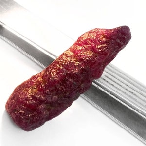 Op de afbeelding: Een ruwe, rode robijn edelsteen met een puntige punt. De robijn is ongeveer 5 cm lang en heeft een natuurlijke, onbewerkte afwerking.