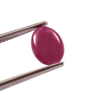 May include: A vibrant pink oval-shaped gemstone held by tweezers. The gemstone is smooth and polished, with a glossy finish.