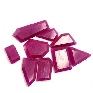 May include: A selection of faceted, deep magenta gemstones in various geometric shapes, including rectangles, trapezoids, and triangles. The gems are arranged on a white background, showcasing their polished surfaces and rich colour.
