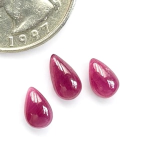 May include: Three teardrop-shaped ruby gemstones, each approximately 1/2 inch long, are arranged in a row on a white surface. A US quarter is shown for size comparison.