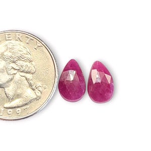 Peut inclure: Deux rubis roses profonds, taillés en forme de goutte, sont présentés à côté d'une pièce de monnaie. Les pierres précieuses ont une surface polie et réfléchissante, sur fond blanc. La pièce est datée de 1997.