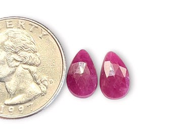 May include: Two faceted, teardrop-shaped, deep pink rubies are displayed next to a quarter coin. The gemstones have a polished, reflective surface and are set against a white background. The quarter coin is dated 1997.