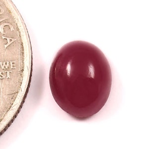 May include: A polished, oval-shaped, deep red gemstone. The gem is smooth and reflective, with a slight sheen. A U.S. quarter coin is next to the gem for size comparison. The gemstone is set against a white background.