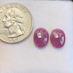 Puede incluir: Dos gemas rosas oscuras, facetadas y de forma ovalada, se muestran junto a una moneda de 25 centavos estadounidense. Las gemas tienen un corte brillante, que refleja la luz. La moneda es plateada y muestra un perfil de George Washington.