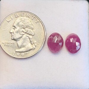 Puede incluir: Dos gemas de rubí ovaladas y facetadas en un engaste de color claro. Una moneda de veinticinco centavos estadounidense está junto a los rubíes para comparar el tamaño. La moneda muestra el perfil de George Washington y el año 1997.