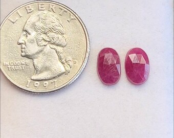 May include: Two oval-shaped, faceted ruby gemstones next to a U.S. quarter coin. The rubies are a deep red color and have a polished, reflective surface. The coin is dated 1997.