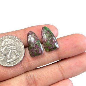 Könnte beinhalten: Zwei grüne und rosafarbene Edelstein-Cabochons mit facettiertem Schliff. Die Steine sind dreieckig geformt und werden in einer Hand zur Größenvergleich gehalten.
