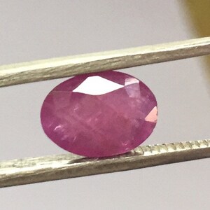May include: A pink oval-shaped gemstone held by tweezers.