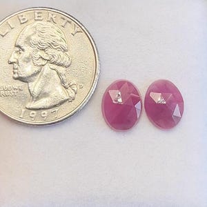 May include: Two oval-shaped, faceted ruby gemstones with a quarter coin for size comparison. The rubies are a deep pinkish-red color and have a sparkling, polished appearance. The coin is a United States quarter from 1997.