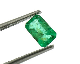 Puede incluir: Una esmeralda verde vibrante, tallada en forma rectangular, está sujeta por unas pinzas.