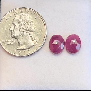 May include: Two faceted, oval-shaped, deep red rubies next to a U.S. quarter. The gemstones have a brilliant cut, reflecting light. The quarter shows George Washington's profile and the year 1997.