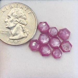 May include: A collection of faceted, hexagonal-shaped, pink gemstones. A U.S. quarter coin is next to the gemstones for size comparison. The coin is dated 1997. The gemstones are arranged on a white surface.