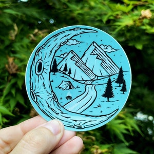 May include: A blue and black sticker with a crescent moon design. The moon is filled with a mountain scene with a river, trees, and a tent.