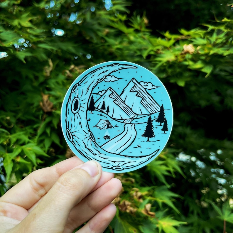 May include: A blue and black sticker with a crescent moon design. The moon is filled with a mountain scene with a river, trees, and a tent.