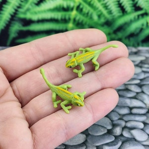 May include: Two small green plastic toy chameleons with yellow stripes. The chameleons are posed on a hand with their tongues extended.