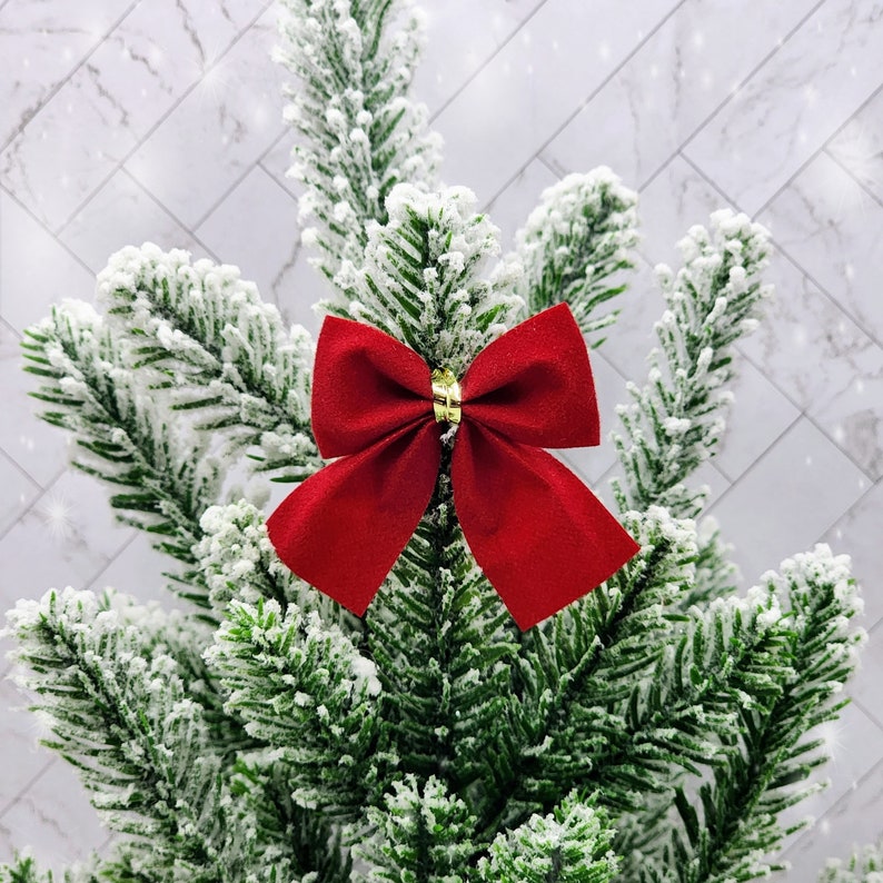 May include: A red velvet bow with a gold centre is attached to a snowy green artificial Christmas tree.