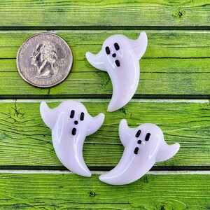 May include: Three white ghost-shaped glass ornaments with black painted eyes and mouths. The ghosts are on a green wooden surface.