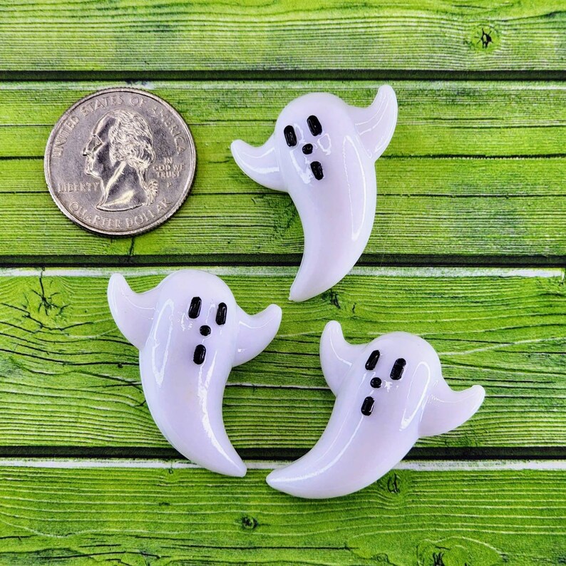 May include: Three white ghost-shaped glass ornaments with black painted eyes and mouths. The ghosts are on a green wooden surface.