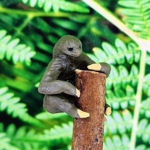 May include: A small, gray sloth figurine with yellow claws is climbing a brown tree branch. The figurine is made of a smooth, textured material and has a realistic appearance.