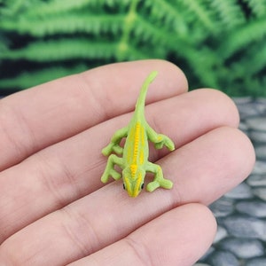 May include: A small green and yellow chameleon figurine. The chameleon is posed with its tail curled up and its legs outstretched.
