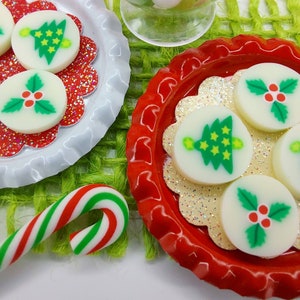 May include: Two red and white plates with a white background and a red rim. Each plate has a white, round, decorative candy with a Christmas design. One candy has a green Christmas tree with yellow stars and a yellow light on top. The other candy has a green holly leaf with red berries. A candy cane is in the foreground.