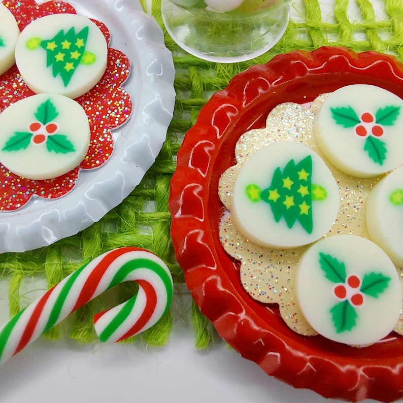 May include: Two red and white plates with a white background and a red rim. Each plate has a white, round, decorative candy with a Christmas design. One candy has a green Christmas tree with yellow stars and a yellow light on top. The other candy has a green holly leaf with red berries. A candy cane is in the foreground.