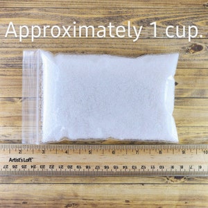 May include: A clear plastic bag filled with white, granulated sugar. The bag is sitting on a wooden surface with a ruler next to it. The ruler reads "Approximately 1 cup."
