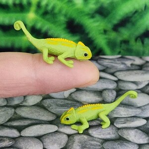 May include: Two small green and yellow toy chameleon figurines. The chameleon on the left is on a finger, while the chameleon on the right is on a bed of small grey rocks.